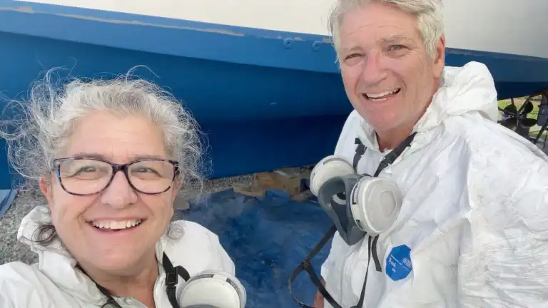 Linda and Matt smiling in protective coveralls beside the freshly restored blue hull of their boat in the boat yard